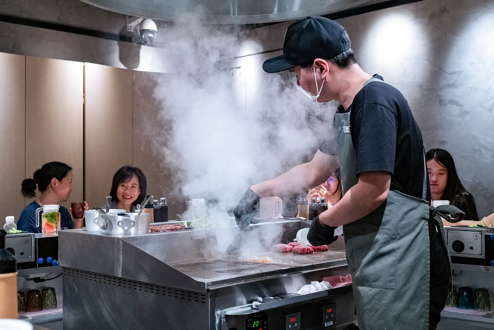 Chef cooks freshly ground Japanese style burgers on a grill while customers look on